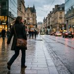 Eine Frau von hinten auf einem nassen Kopfsteinpflaster in einer belebten Stadtstraße, umgeben von historischen Gebäuden, Straßenbahn, Autos und Passanten bei regnerischem Abenddämmerungslicht. Die Straße führt als führende Linie in die Bildtiefe.